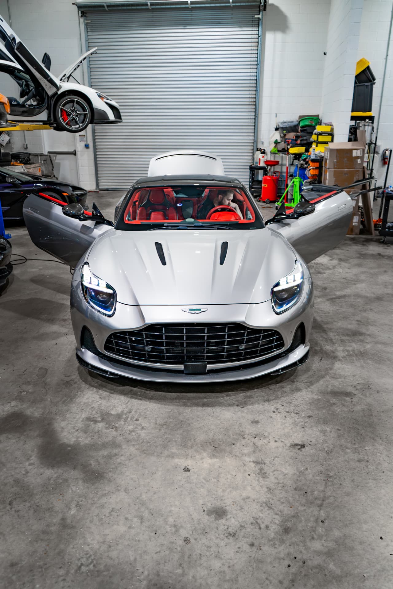 Silver Aston Martin DB12 head-on in the March Motorsports workshop with both doors open and red interior visible