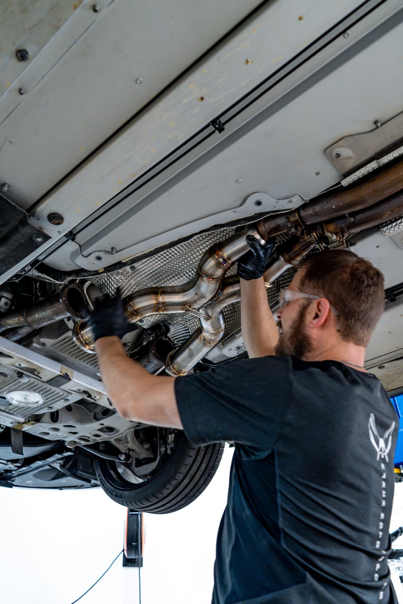 Technician fitting a stainless X-pipe exhaust under an Aston Martin Vanquish at March Motorsports